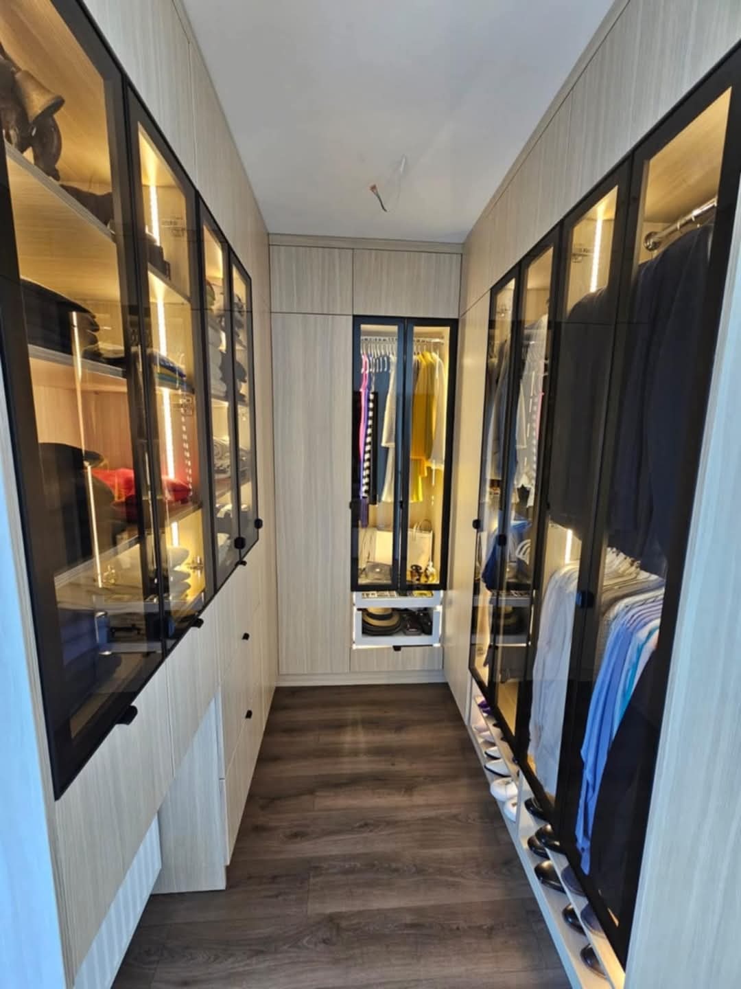 Nowoczesna garderoba na wymiar z przeszklonymi frontami w czarnych ramach i oświetleniem LED, ukazująca uporządkowane ubrania i akcesoria.