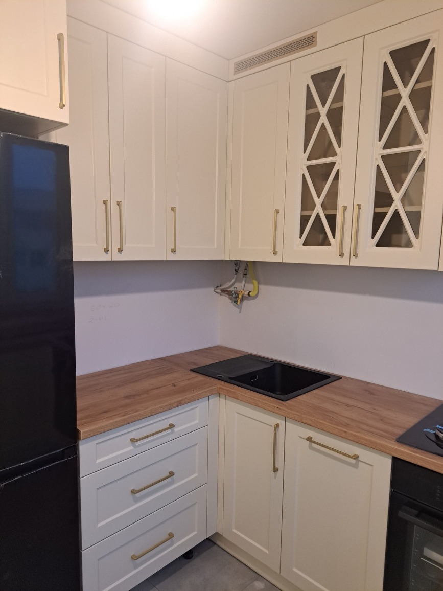 Jasna kuchnia z drewnianym blatem i złotymi uchwytami. Górne szafki z przeszkleniami w geometryczne wzory. Widoczny fragment czarnej lodówki.