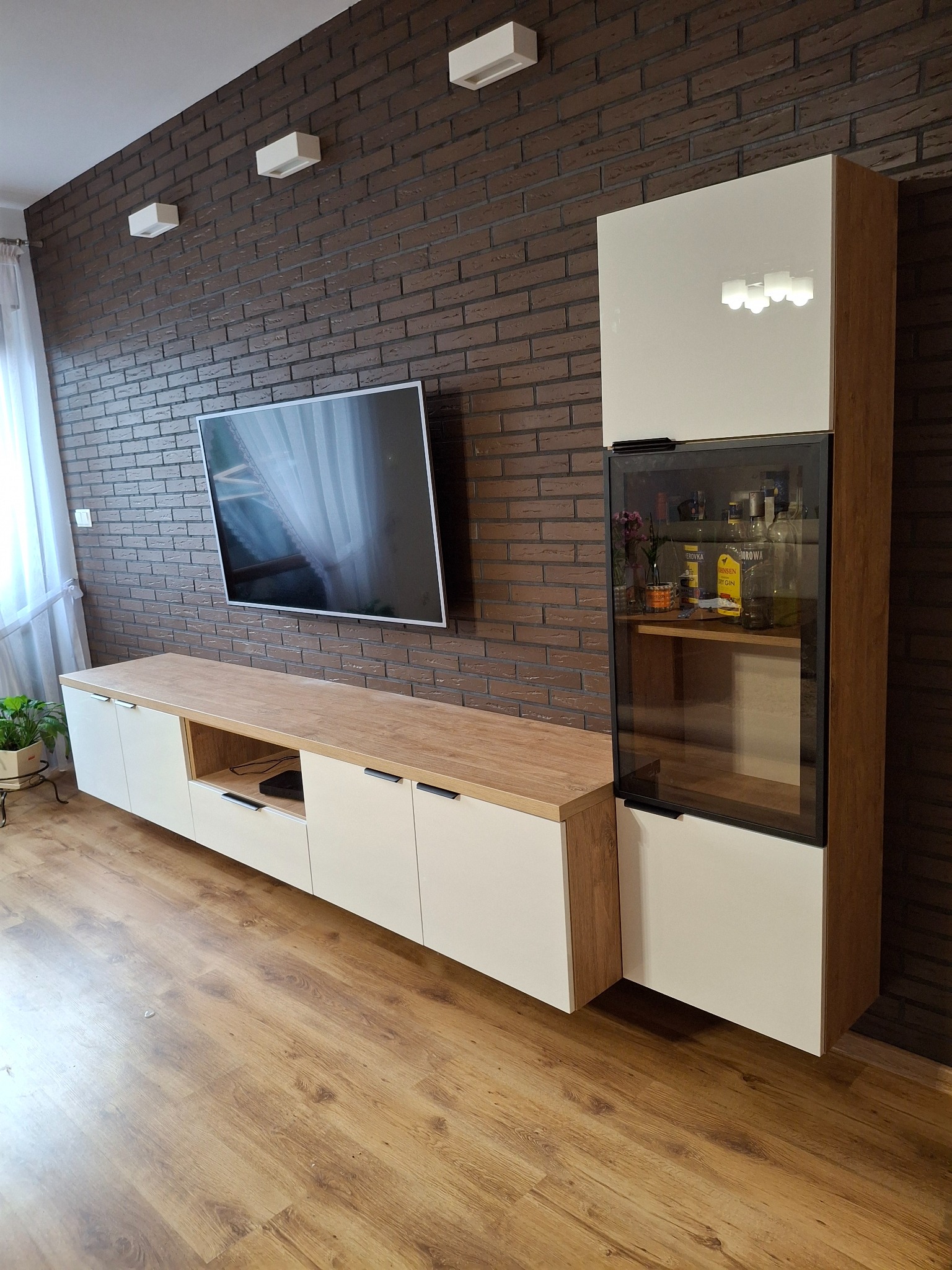 Nowoczesny zestaw mebli RTV z witryną na tle ściany z cegły. Białe fronty, drewniany blat, czarne uchwyty. W witrynie szkło i oświetlenie.