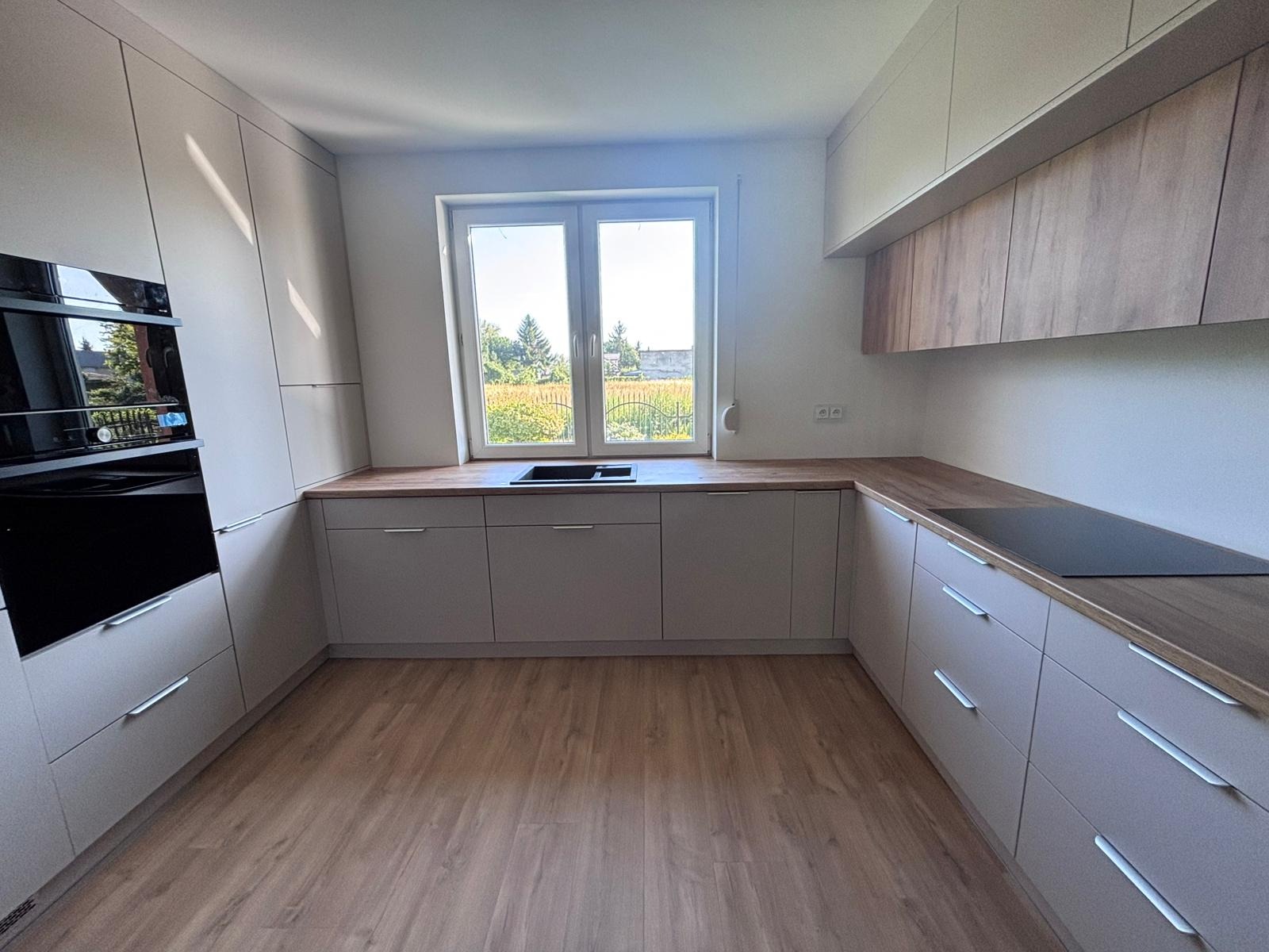 Jasna kuchnia w kształcie litery U z drewnianym blatem i widokiem na zieleń za oknem. Minimalistyczny design, fronty bez uchwytów, płyta indukcyjna.