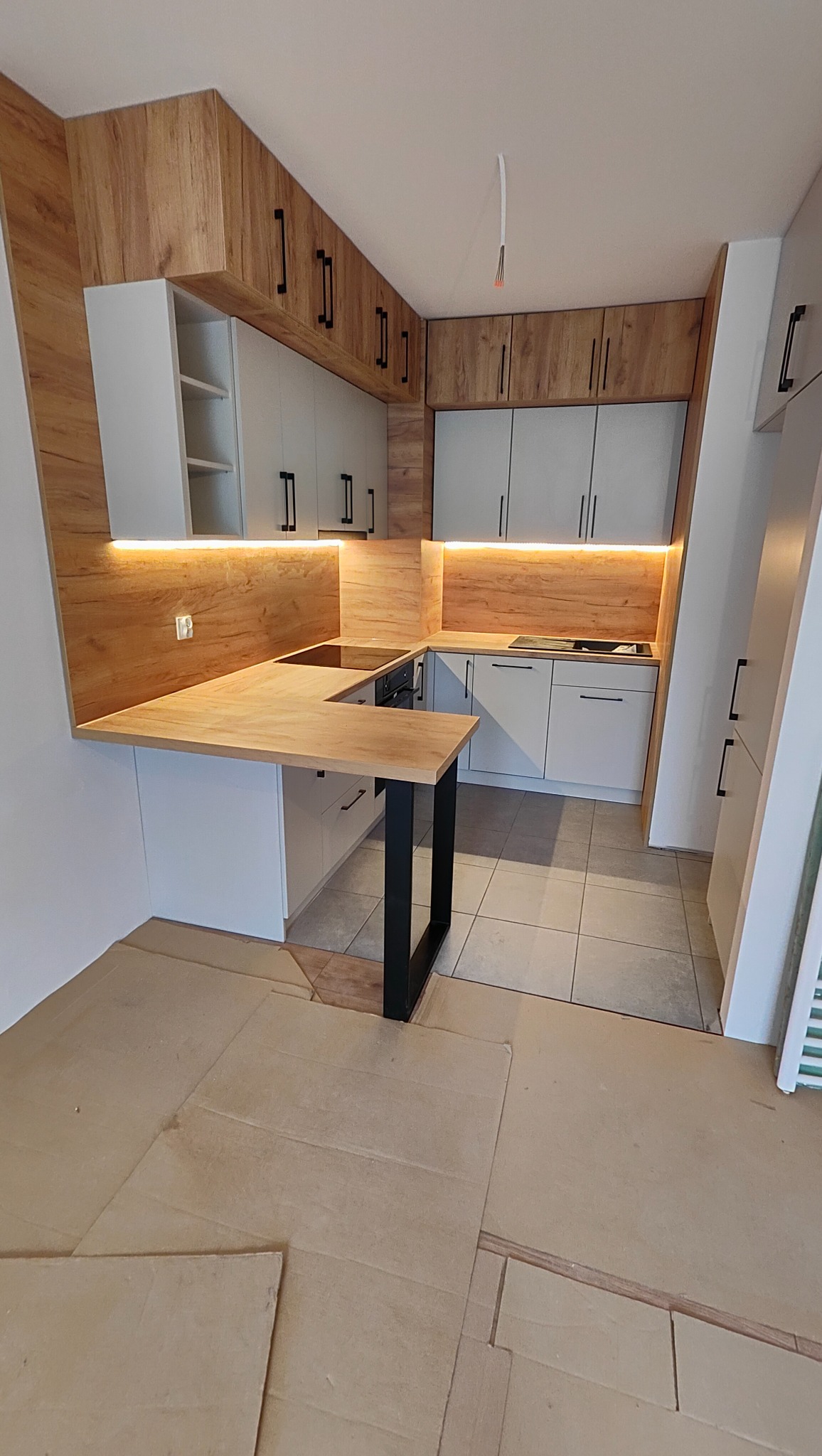 Nowoczesna kuchnia z drewnianymi akcentami i białymi frontami, oświetlona listwami LED, z blatem w kształcie litery U i czarną nogą stołu. Podłoga zabezpieczona kartonem.