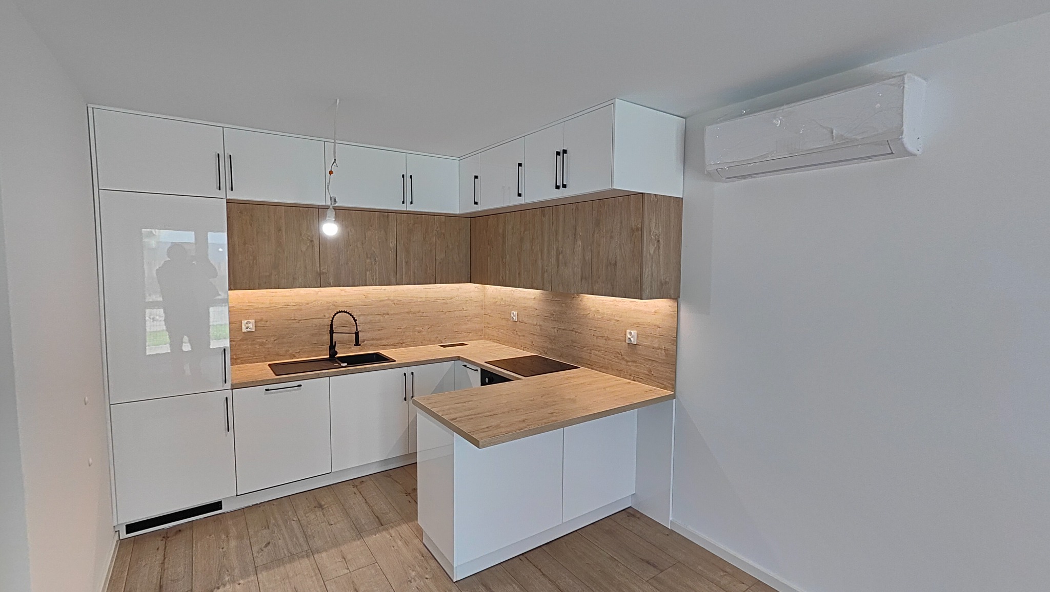 Nowoczesna kuchnia na wymiar z białymi frontami i drewnopodobnym blatem, oświetlenie LED pod szafkami, czarna armatura, klimatyzator na ścianie.
