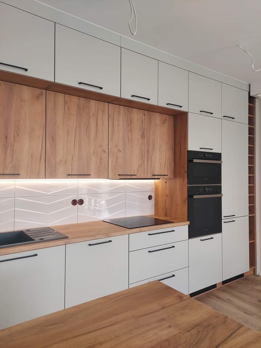 Nowoczesna kuchnia z białymi i drewnianymi szafkami, czarnymi uchwytami i sprzętem AGD. Uwagę przykuwają płytki z subtelnym wzorem jodełki nad blatem.