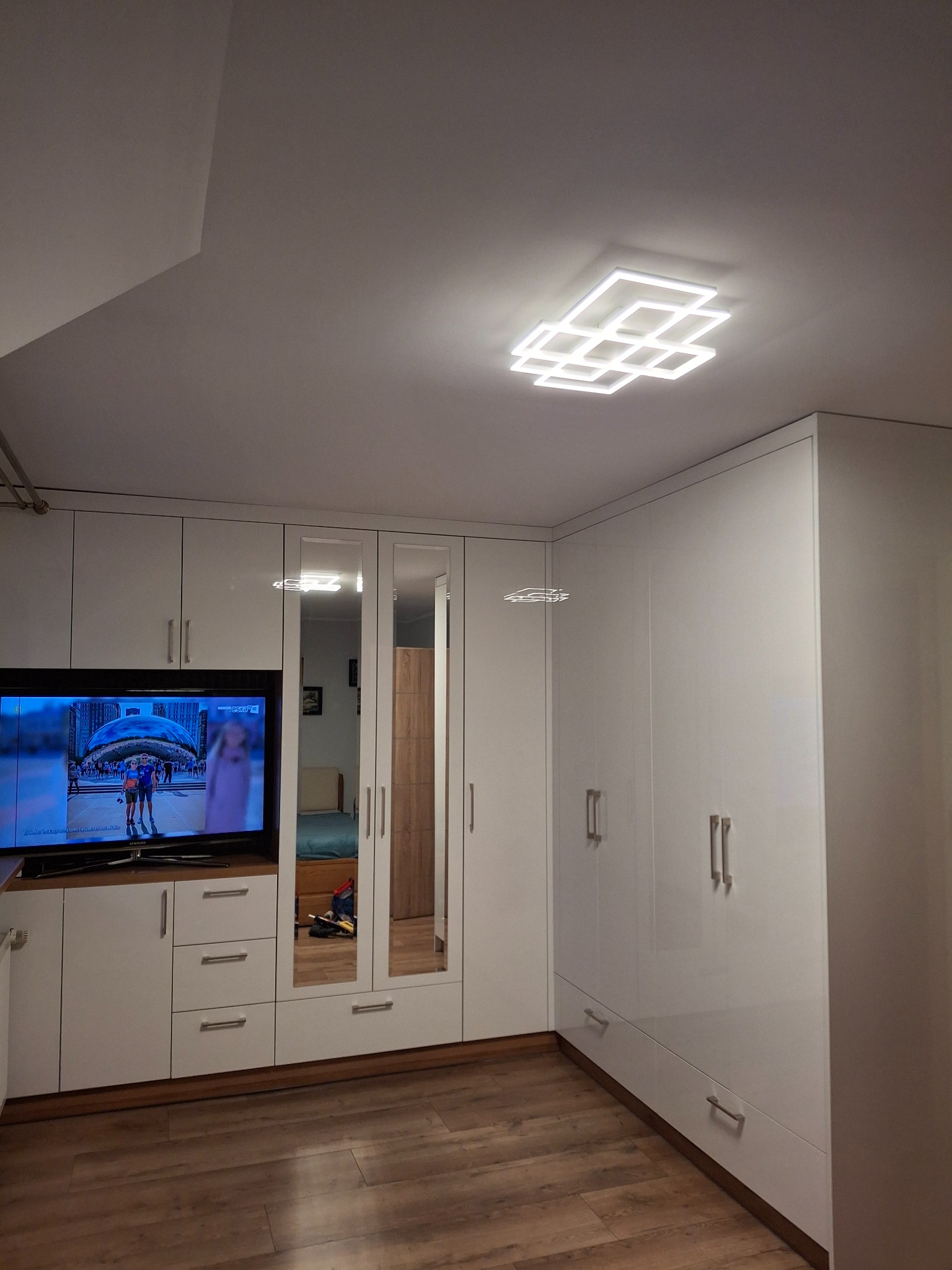 Biała szafa wnękowa z lustrami i telewizorem wbudowanym w część meblową, oświetlenie sufitowe w kształcie kwadratów, drewniana podłoga.