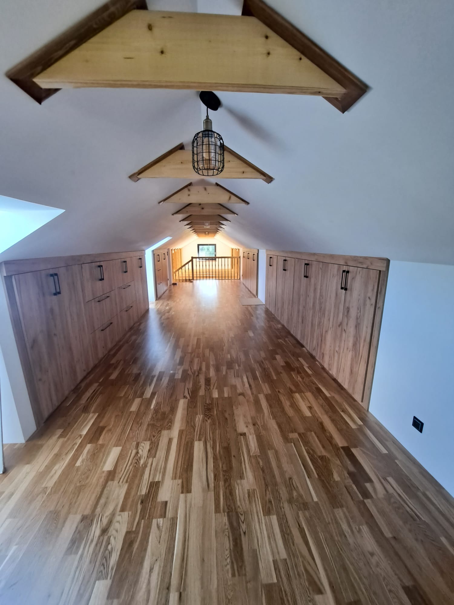 Długi korytarz na poddaszu z drewnianą podłogą i zabudowanymi szafami wnękowymi po obu stronach, oświetlony wiszącą lampą w stylu industrialnym.
