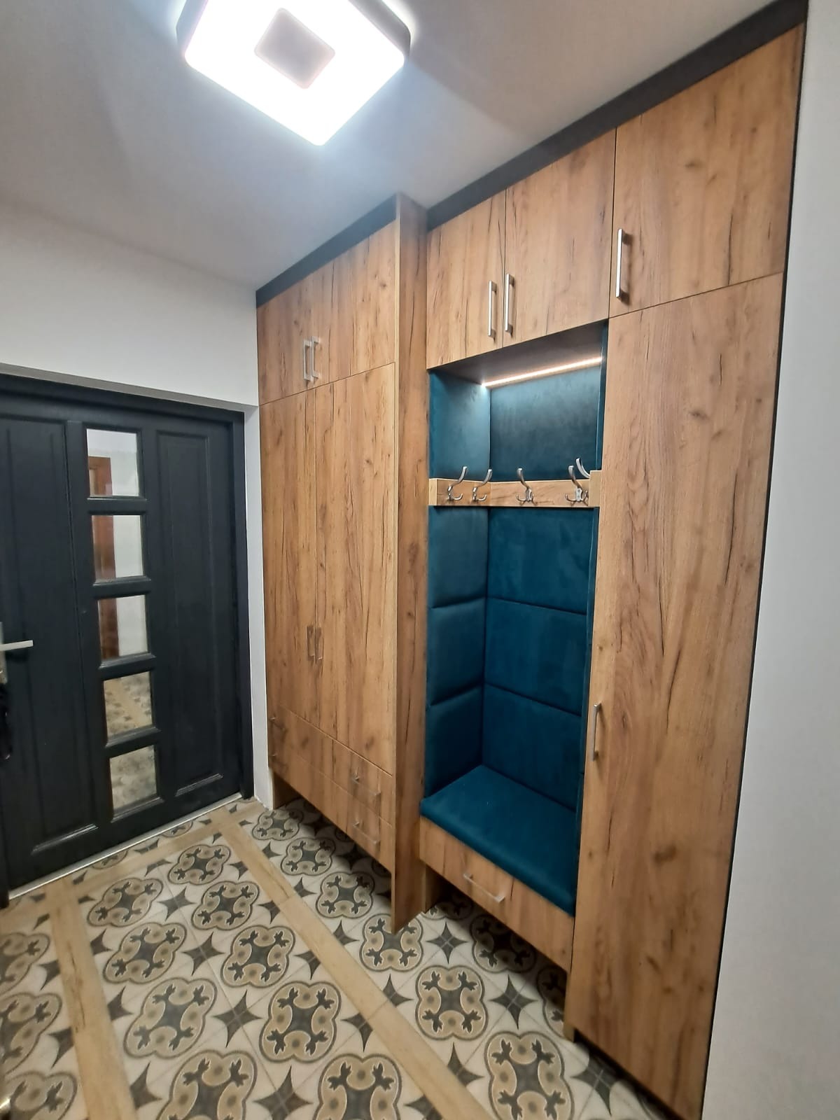 Funkcjonalna szafa w przedpokoju z siedziskiem obitym turkusowym materiałem i wieszakami, drewnopodobne fronty, oświetlenie LED, czarne wykończenia.