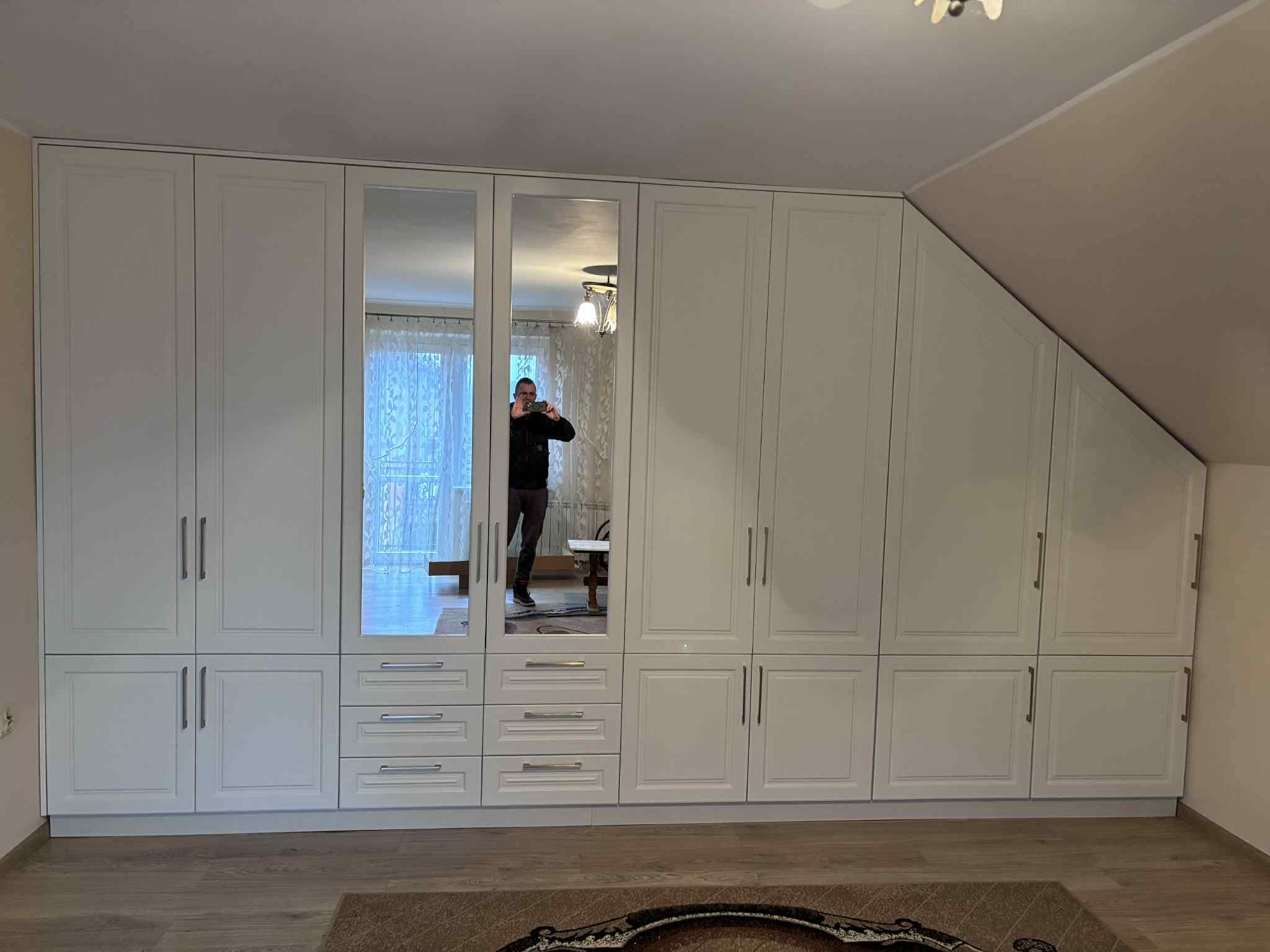 Biała szafa wnękowa z lustrami, dopasowana do skosu dachu, z widocznym odbiciem osoby robiącej zdjęcie. Klasyczny design, srebrne uchwyty, drewniana podłoga.