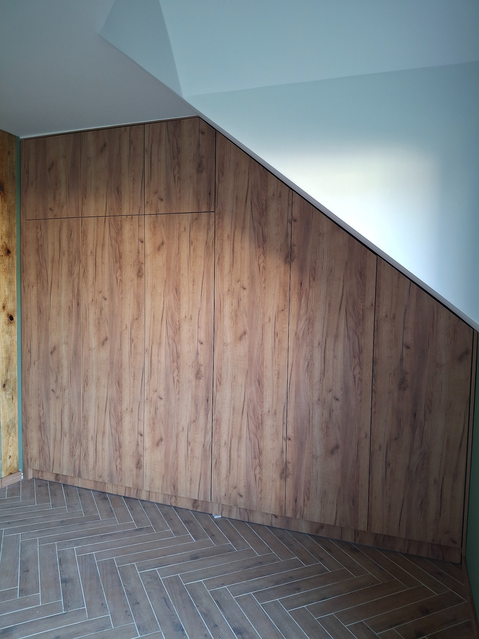 Szafa wnękowa z drewnopodobnymi frontami, dopasowana do skosu poddasza, z widoczną podłogą ułożoną w jodełkę i fragmentem zielonej ściany.