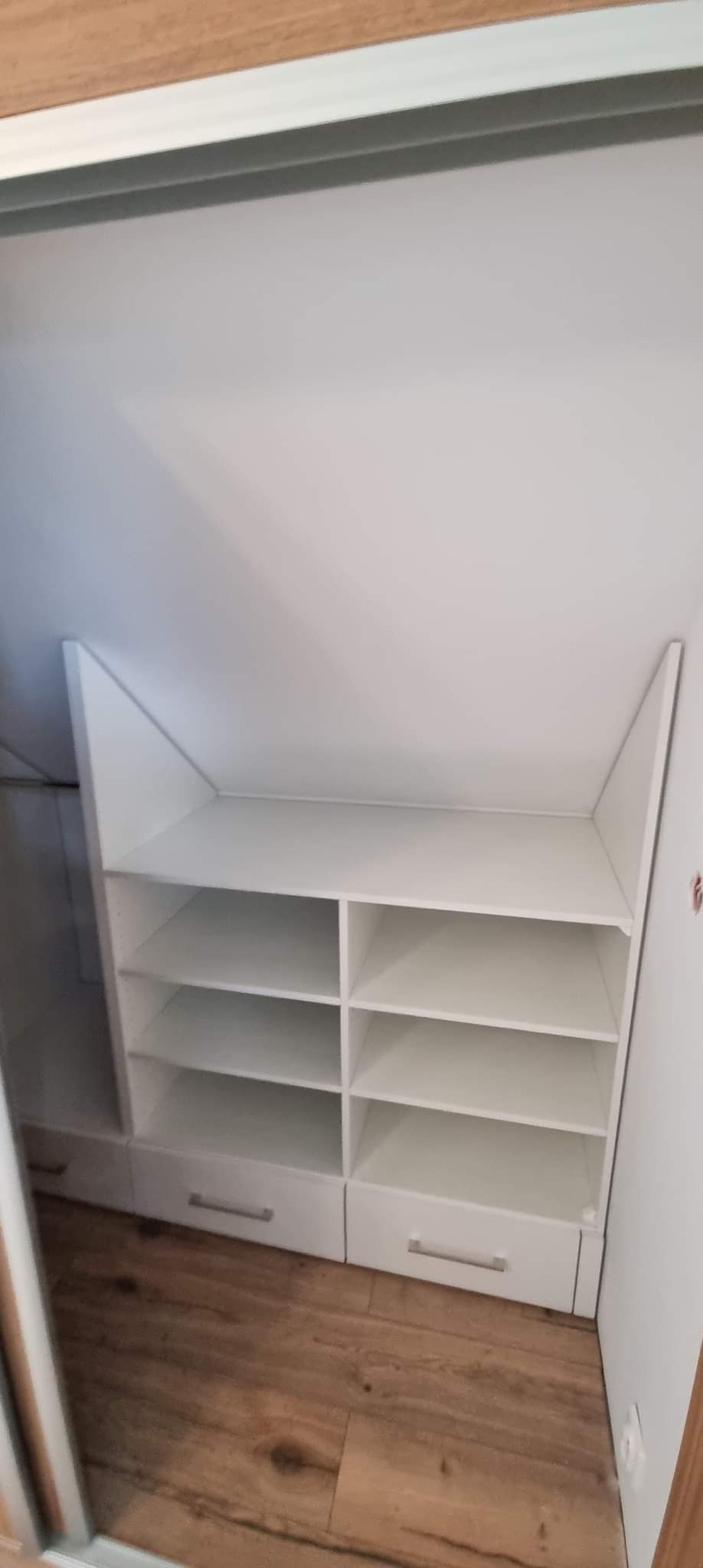 Biała szafa wnękowa z półkami i szufladami, dopasowana do skosu dachu, widok z wnętrza szafy, drewniana podłoga.
