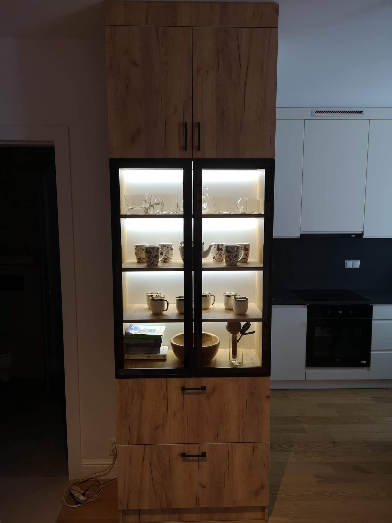 Wysoka witryna z oświetleniem LED, drewnopodobne fronty z czarnymi ramami, szklane półki z naczyniami. W tle zabudowa kuchenna w bieli i czerni.