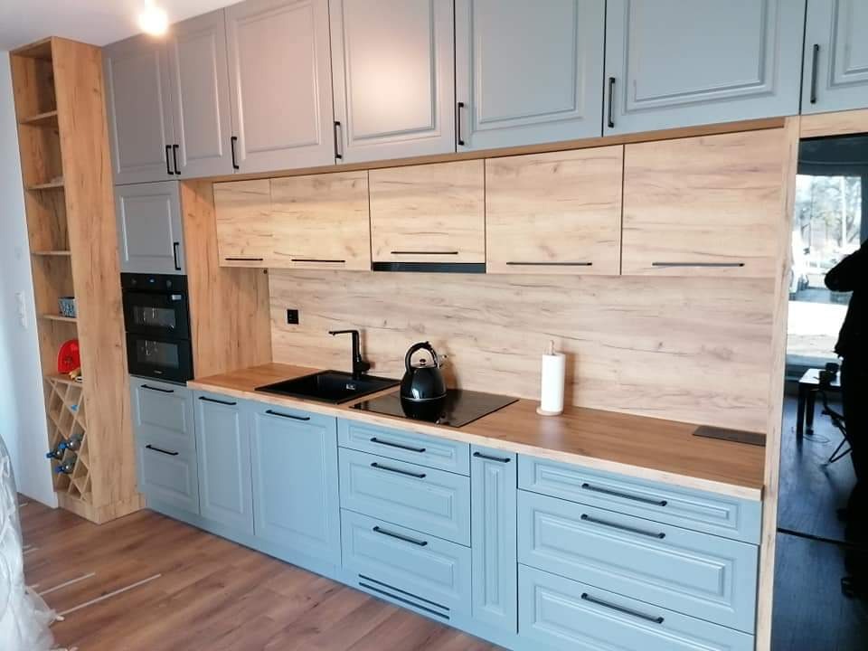 Niebiesko-szara kuchnia z drewnianymi akcentami: szafki górne, blat i regał na wino. Czarny zlew i czajnik. Minimalistyczne uchwyty. Nowoczesny design.