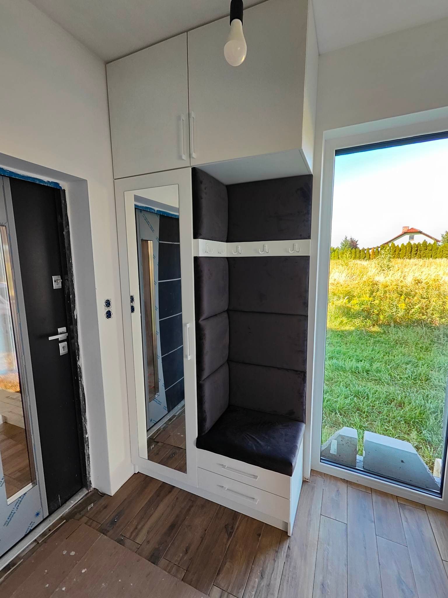 Funkcjonalna zabudowa przedpokoju z siedziskiem i lustrem, białe szafki wiszące, tapicerowany panel ścienny w kolorze grafitowym, widok na zewnątrz przez okno.