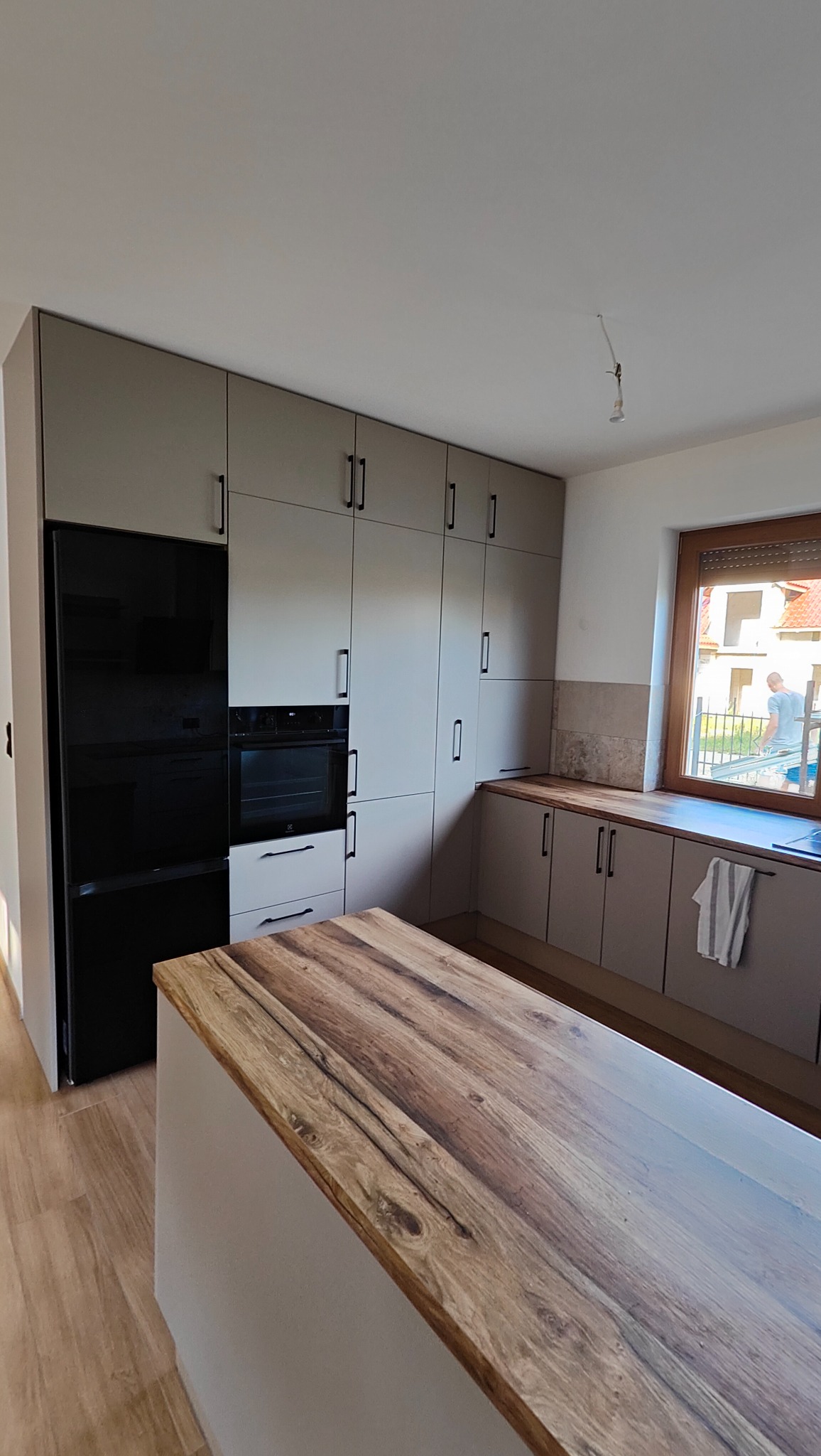 Jasnoszara zabudowa kuchenna z czarnym sprzętem AGD i drewnianym blatem wyspy. Minimalistyczny design, pionowe uchwyty, widok na okno z zewnątrz.