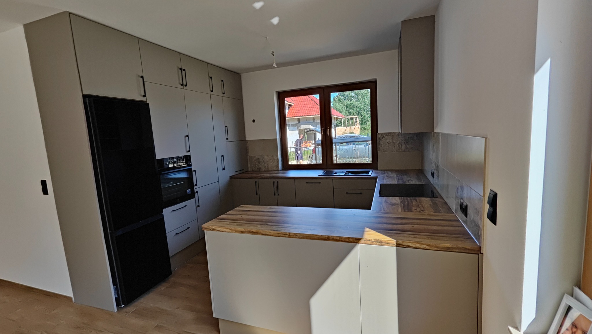 Jasnoszara kuchnia z drewnianym blatem i czarną lodówką. Widok na okno z widokiem na dom z czerwoną dachówką. Nowoczesny design, minimalistyczne uchwyty.