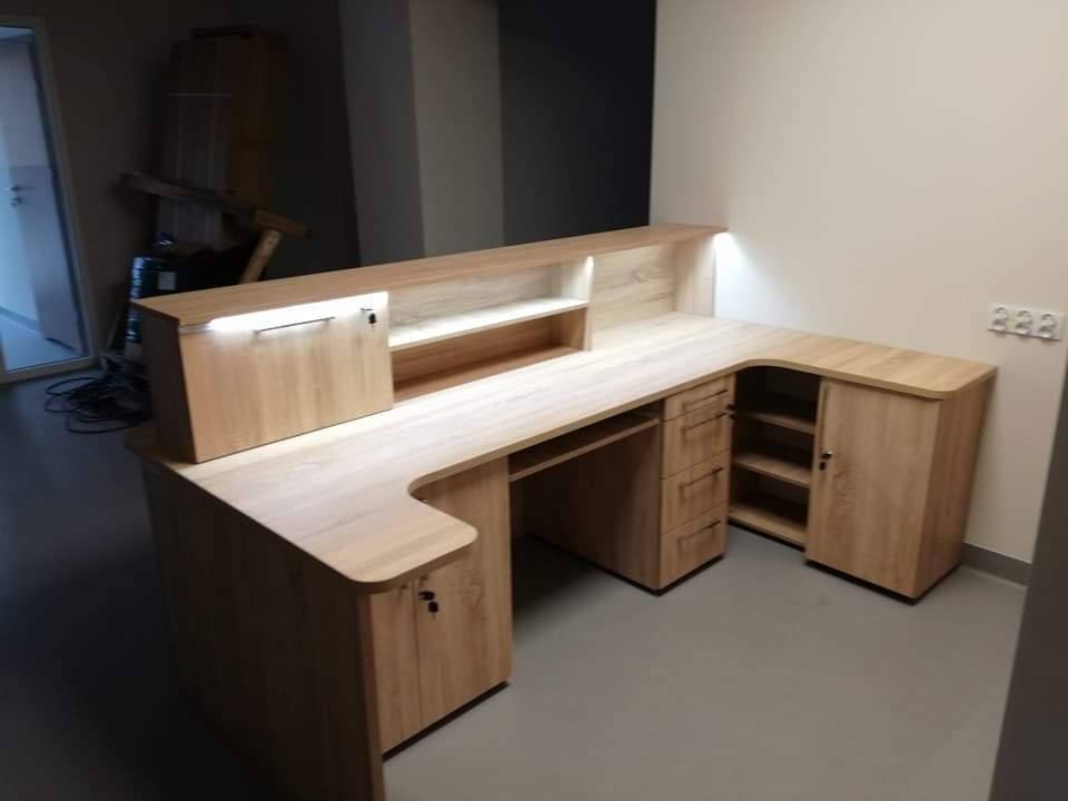 Jasna, narożna lada recepcyjna z oświetleniem LED, wykonana z drewna w odcieniach beżu. Widoczne szafki, półki i przestrzeń na komputer. Nowoczesny design.