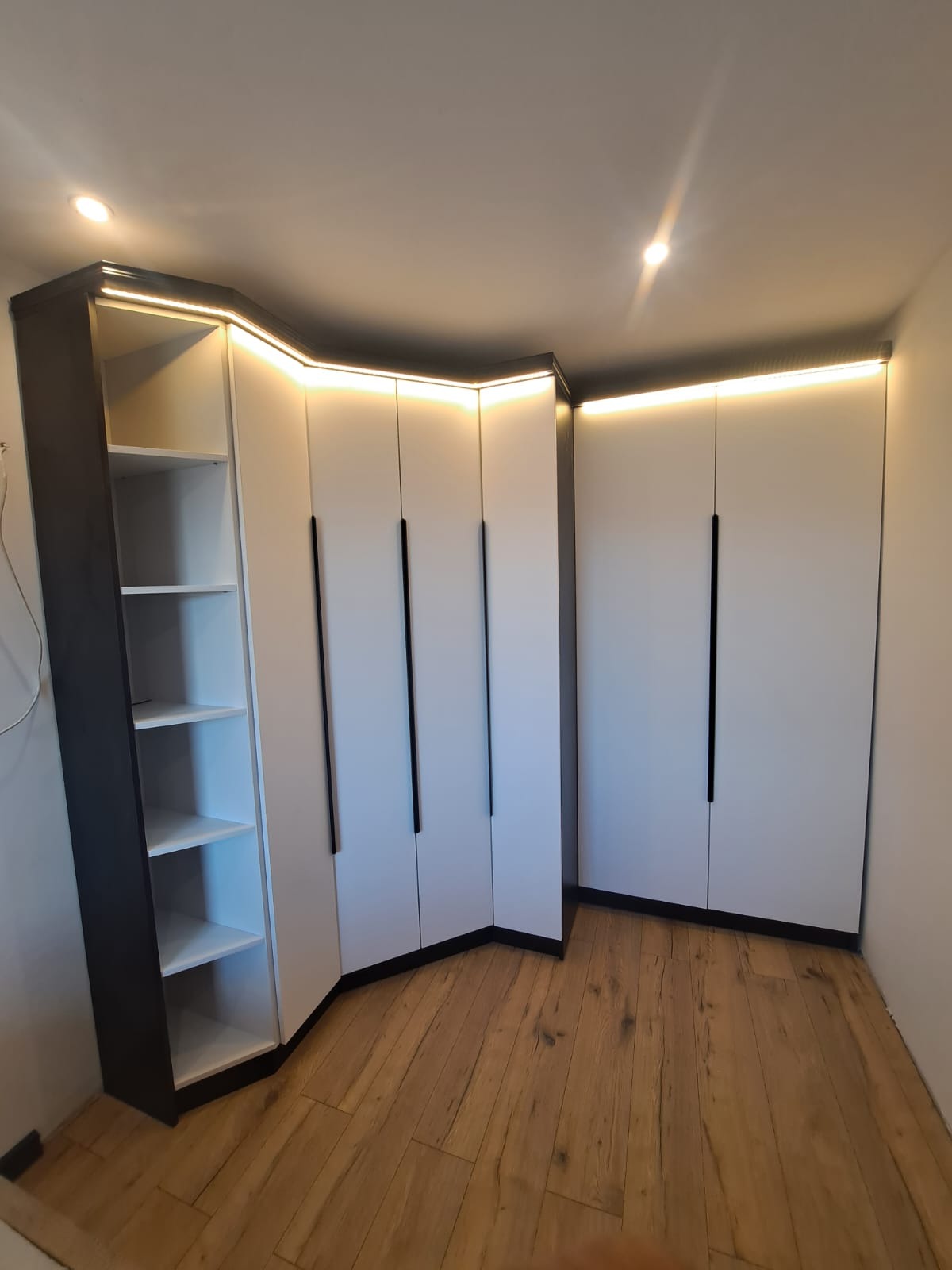 Funkcjonalna szafa wnękowa narożna z oświetleniem LED, białe fronty z czarnymi uchwytami, drewniana podłoga, widok z dołu.