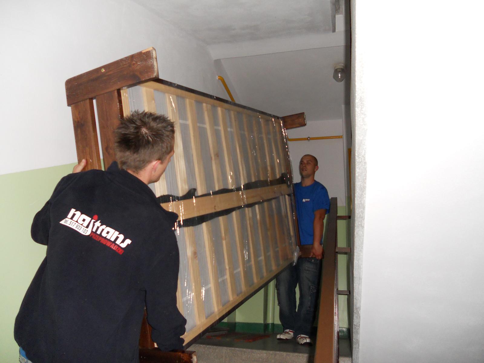 Dwóch mężczyzn w trakcie wnoszenia dużego, zabezpieczonego folią i taśmą drewnianego stelaża po schodach w klatce schodowej. Jeden z nich ma na plecach bluzę z logo firmy transportowej.