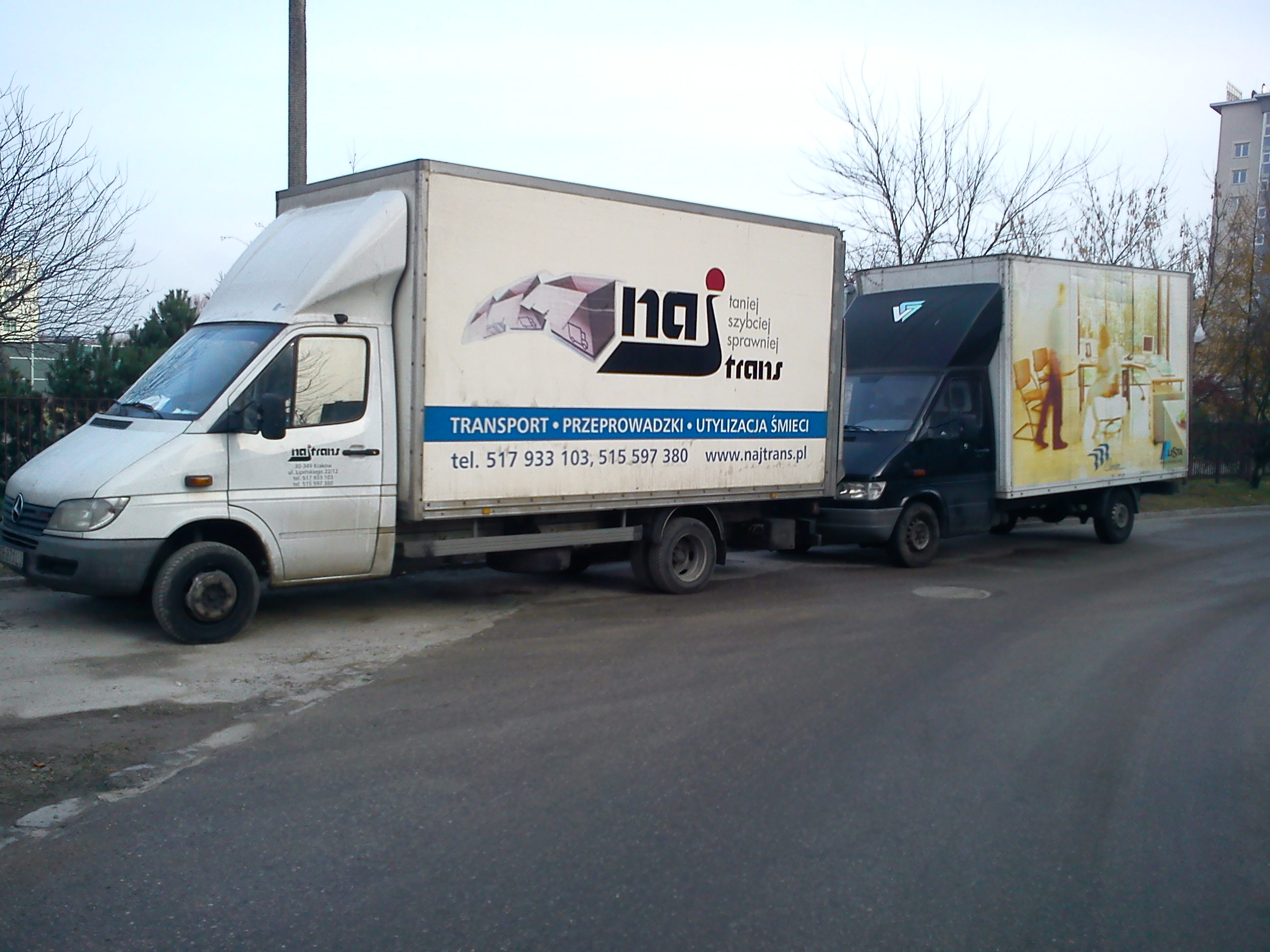 Dwa samochody dostawcze firmy przeprowadzkowej Najtrans zaparkowane na ulicy; biały samochód z reklamą firmy z numerami telefonów i hasłem 'taniej, szybciej, sprawniej trans', za nim ciemny...