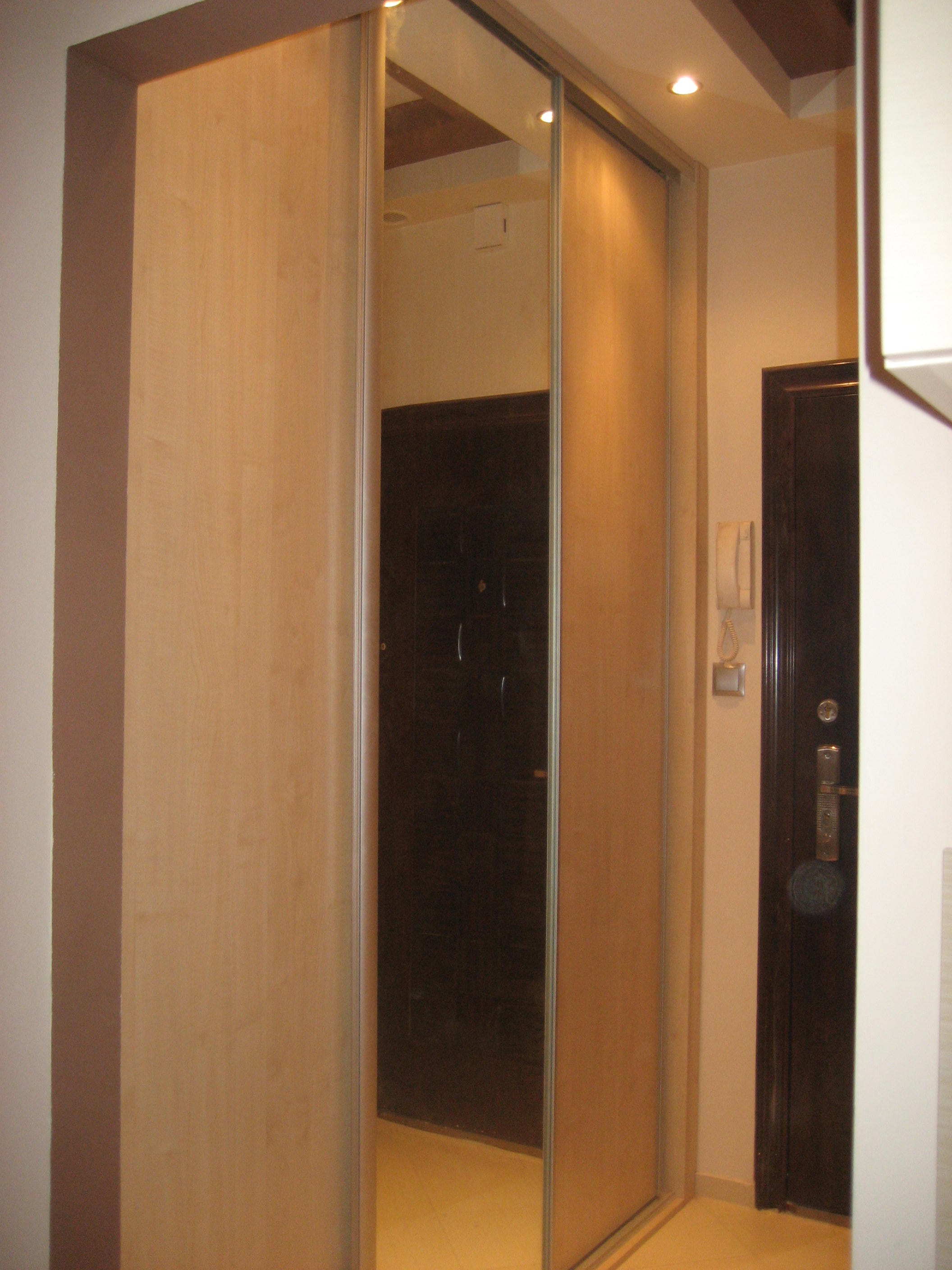 Wnętrze przedpokoju z jasną szafą przesuwną z lustrem i ciemnymi drzwiami wejściowymi z domofonem.