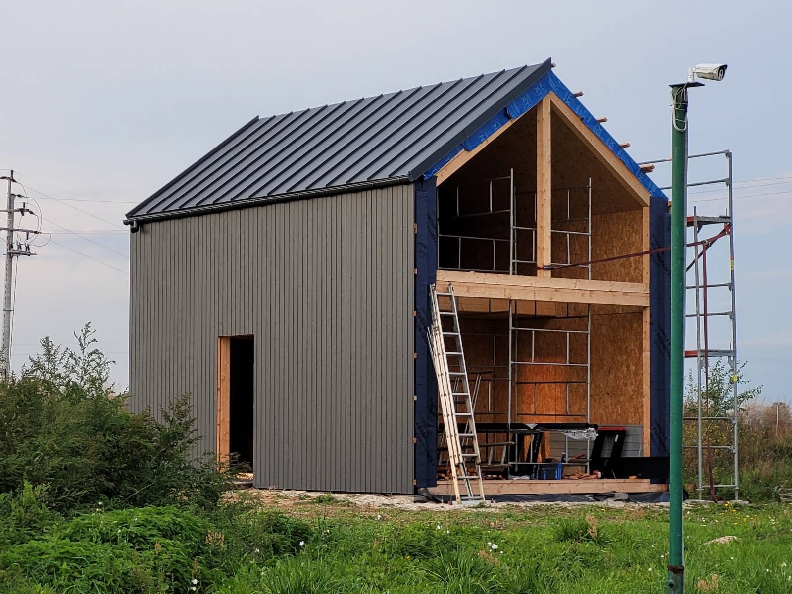 Wznoszenie dwukondygnacyjnego domu w technologii szkieletowej: szara elewacja z pionowym deskowaniem, czarny dach, widoczne rusztowania i drabina przy ścianie bocznej, otwarty szkielet...