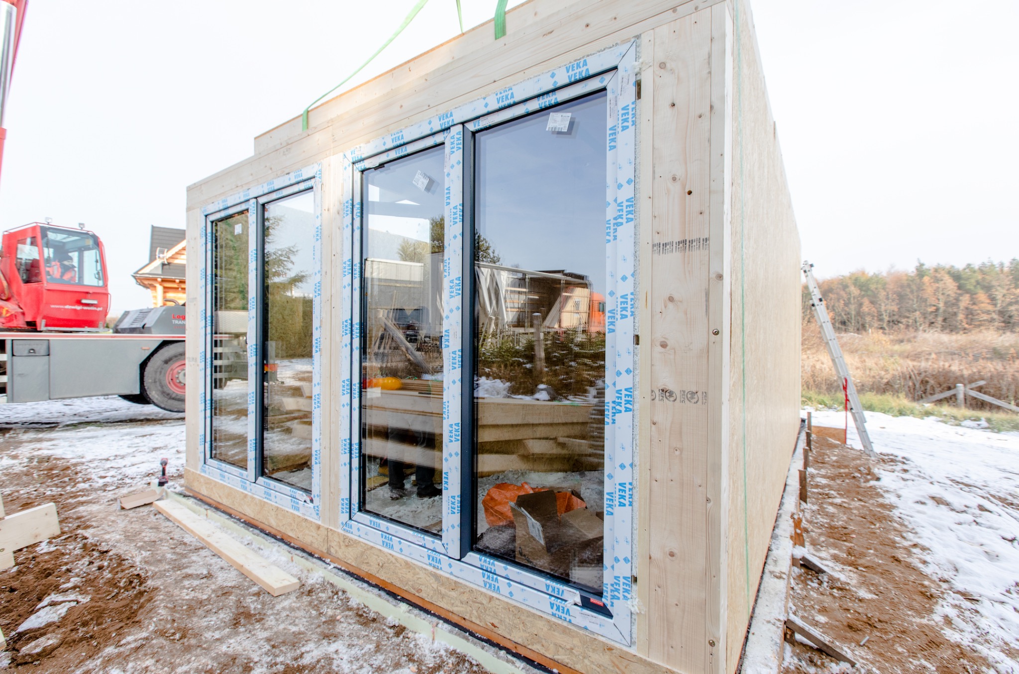 Moduł domu z zamontowanymi oknami VEKA, podnoszony dźwigiem na placu budowy w zimowej scenerii.