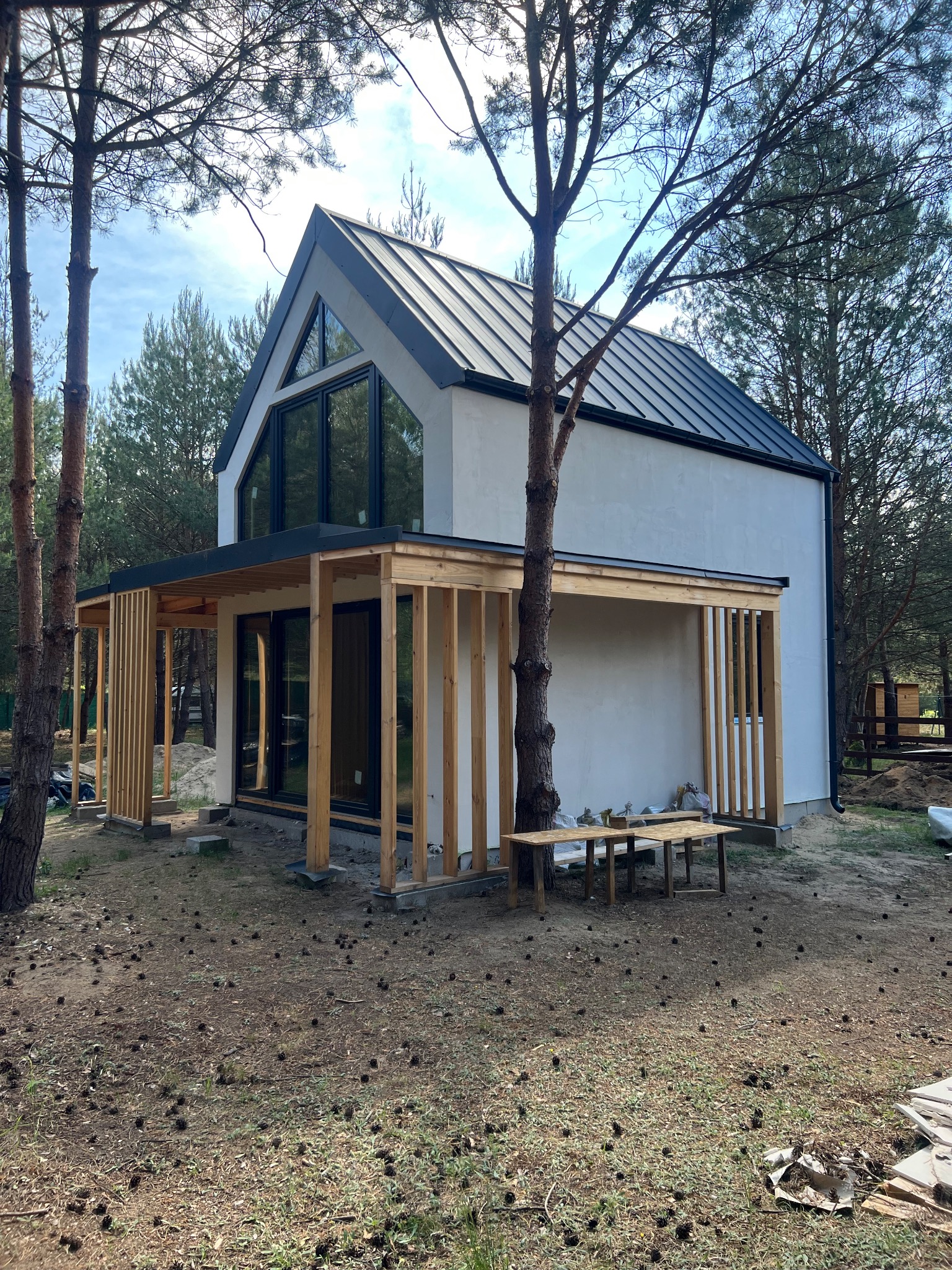 Nowoczesny dom z płaskim dachem i dużymi oknami, otoczony lasem sosnowym, z drewnianą konstrukcją werandy w trakcie budowy i prowizorycznymi stołami na zewnątrz.