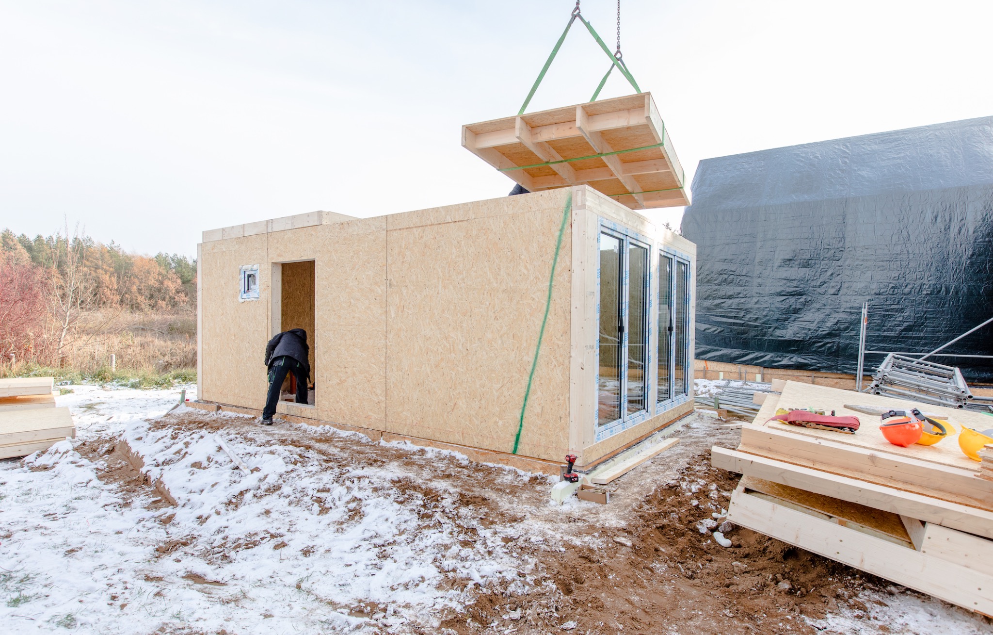 Montaż domu modułowego: pracownik w czarnej kurtce montuje ścianę, podczas gdy dźwig opuszcza prefabrykowany strop na budynek z dużymi oknami, w tle widoczna czarna plandeka.