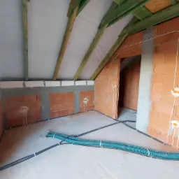 Wnętrze w stanie surowym z widocznymi przewodami elektrycznymi w peszlach i wyprowadzonymi kablami ze ścian, przygotowane do dalszych prac wykończeniowych.