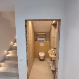 Widok na wykończoną łazienkę z toaletą podwieszaną, umywalką nablatową na drewnianym blacie i lustrem, przez surowy otwór drzwiowy w trakcie remontu, obok widoczne schody z podświetleniem.