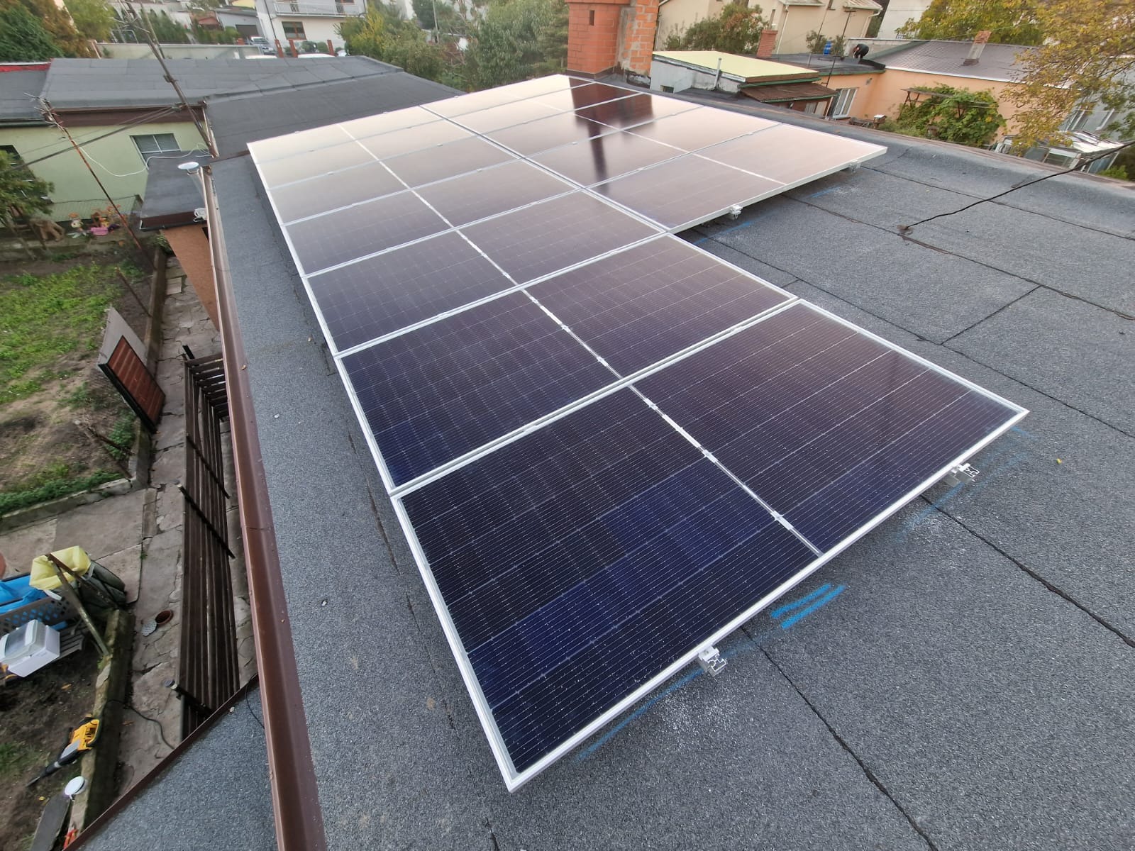 Panele słoneczne na płaskim dachu pokrytym papą, niebieskie ślady montażowe, widok z góry.