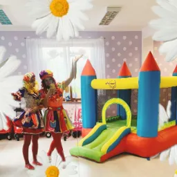 Dwie osoby przebrane w kolorowe stroje stoją obok dmuchanego zamku w pokoju z dekoracjami w kształcie kwiatów; jedna osoba robi selfie telefonem.