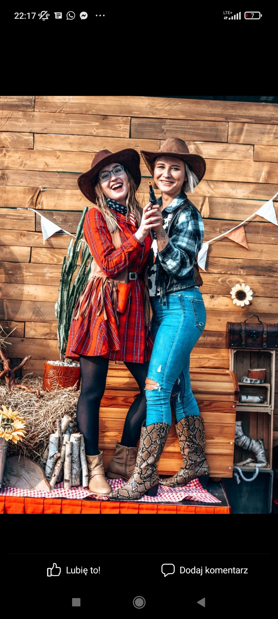 Dwie uśmiechnięte kobiety w strojach kowbojskich, w kapeluszach i butach ze wzorem skóry węża, pozują z pistoletem na tle drewnianej ścianki z dekoracjami w stylu western, w tym kaktus, snopki...