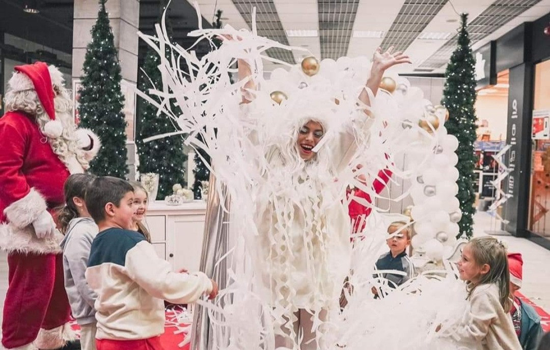 Kobieta w białym stroju z papierowymi serpentynami w centrum uwagi, otoczona uśmiechniętymi dziećmi i postacią Świętego Mikołaja na tle świątecznych dekoracji w galerii handlowej.