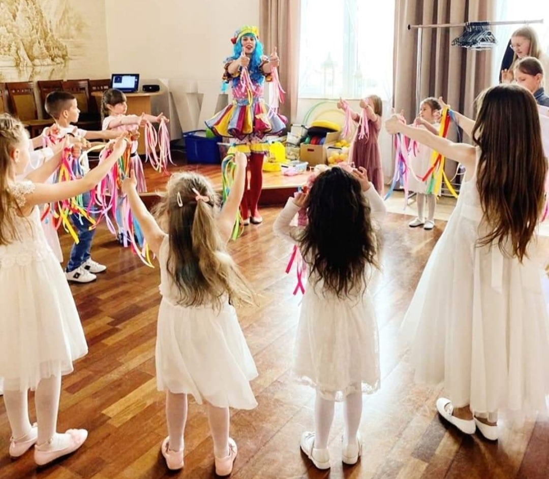 Dzieci w białych sukienkach stoją w kręgu, trzymając kolorowe wstążki, podczas gdy osoba w barwnym kostiumie i niebieskiej peruce pokazuje kciuk w górę.