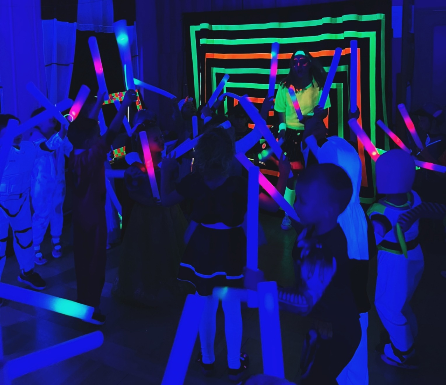 Dzieci bawią się świecącymi pałeczkami w ciemnym pomieszczeniu z neonową dekoracją, niektóre ubrane w kostiumy.