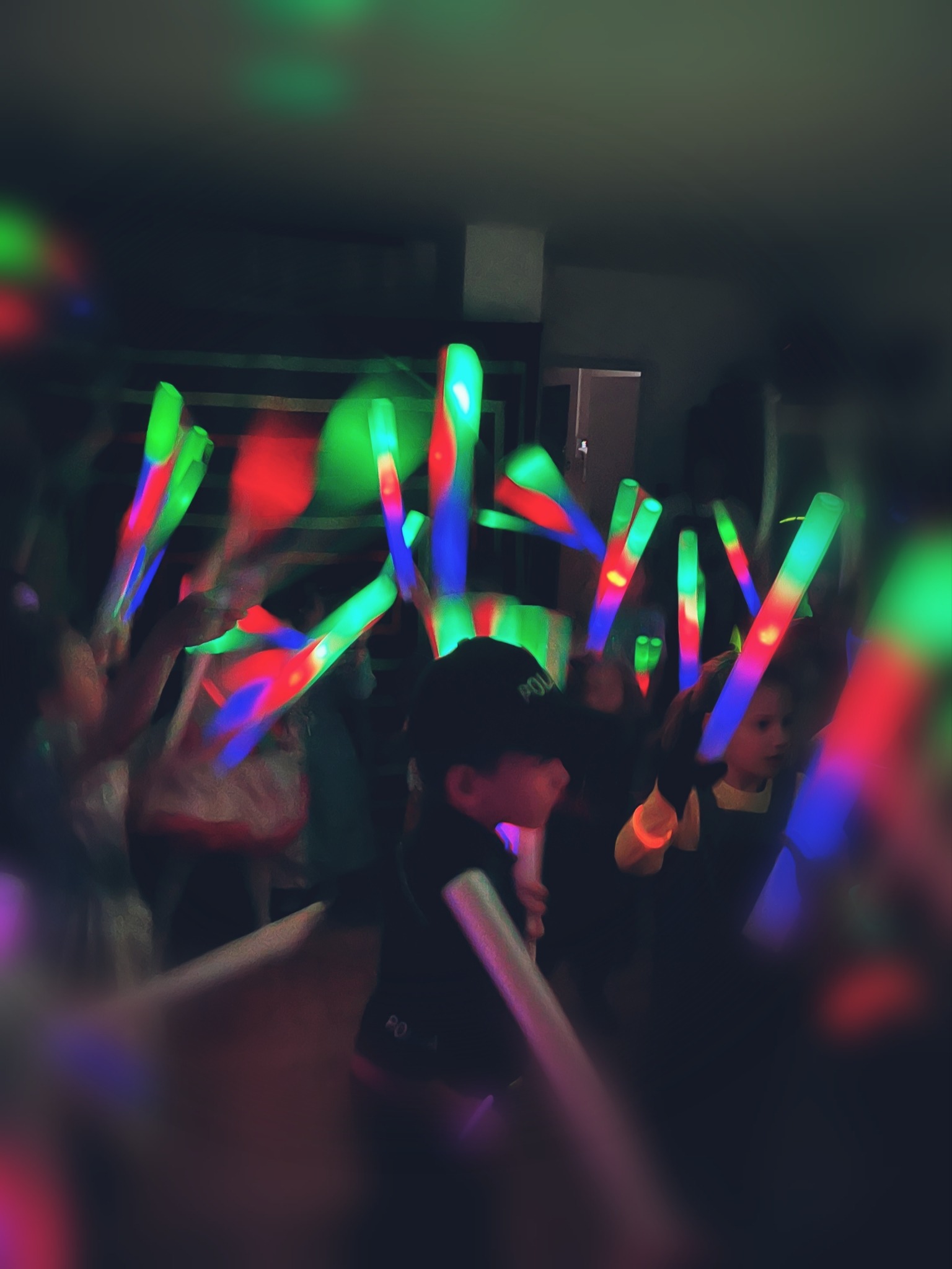 Dzieci w ciemnym pomieszczeniu machają kolorowymi, świecącymi pałeczkami podczas zabawy; chłopiec w czapce z daszkiem na pierwszym planie.