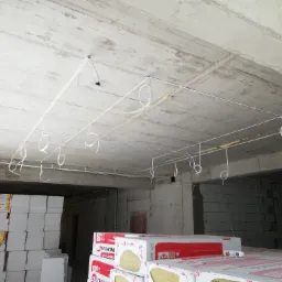 Surowy betonowy sufit z rozprowadzonymi przewodami elektrycznymi w białych peszlach, widoczne puszki instalacyjne, w tle materiały izolacyjne na budowie.