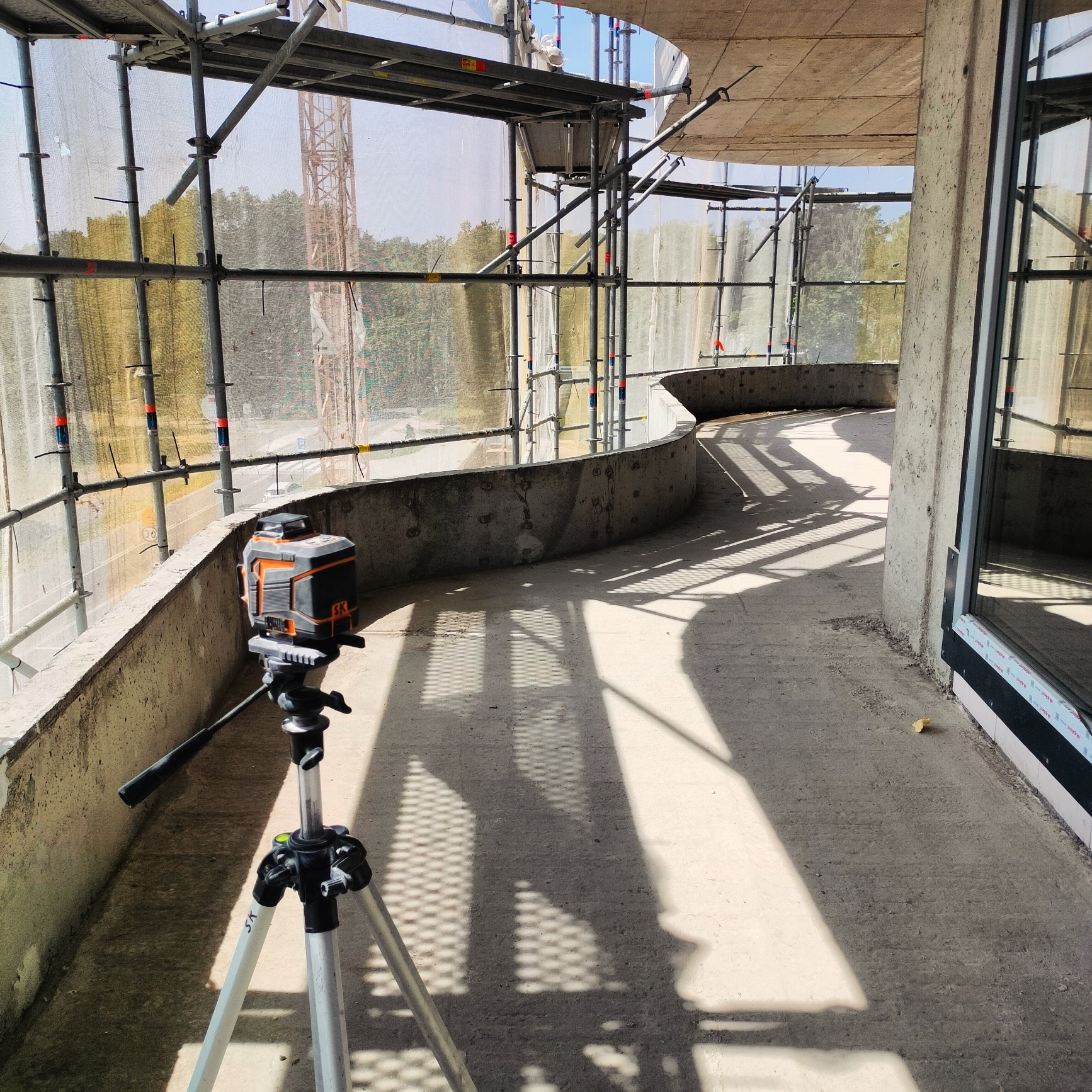Laser krzyżowy na statywie ustawiony na betonowym balkonie w budynku w trakcie budowy, w tle widoczne rusztowania i siatka ochronna.