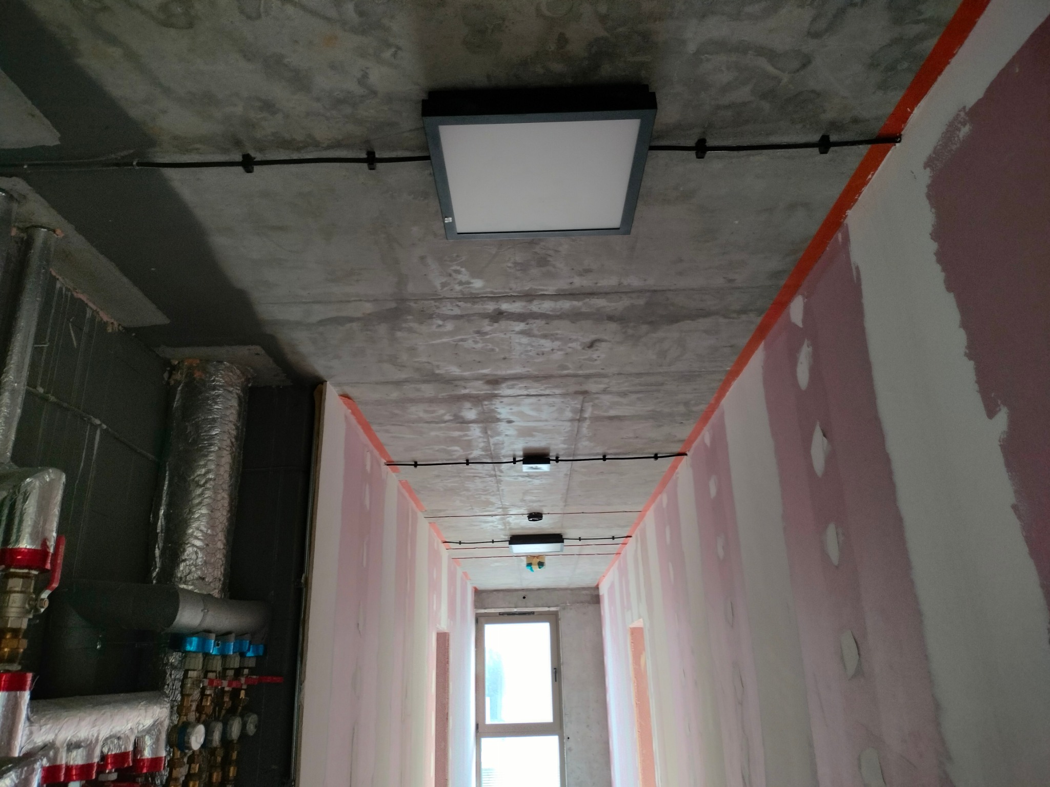 Wnętrze korytarza w stanie surowym z widocznymi przewodami elektrycznymi i zamontowanymi panelami LED na suficie, z instalacją grzewczą po lewej stronie.