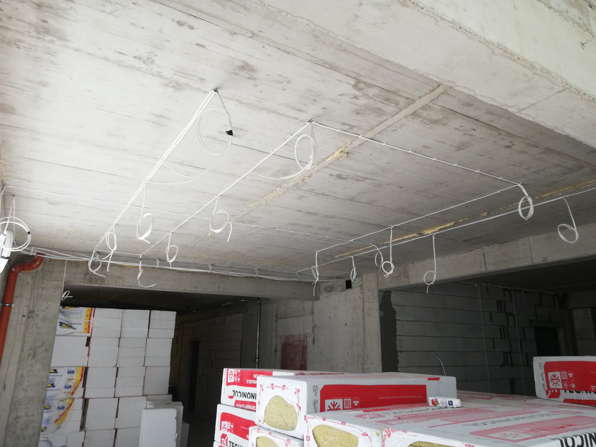 Surowy betonowy sufit z rozprowadzonymi przewodami elektrycznymi w białych peszlach, widoczne puszki instalacyjne, w tle materiały izolacyjne na budowie.