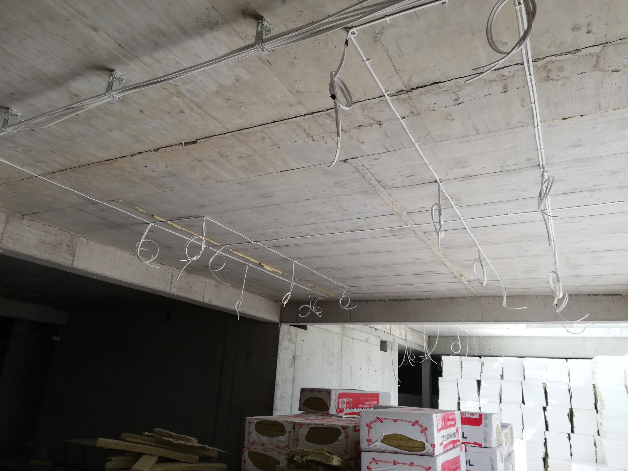 Niedokończona instalacja elektryczna na surowym betonowym suficie, przewody w białych rurkach mocowane do sufitu za pomocą metalowych uchwytów, zwisające pętle kabli, w tle materiały izolacyjne...