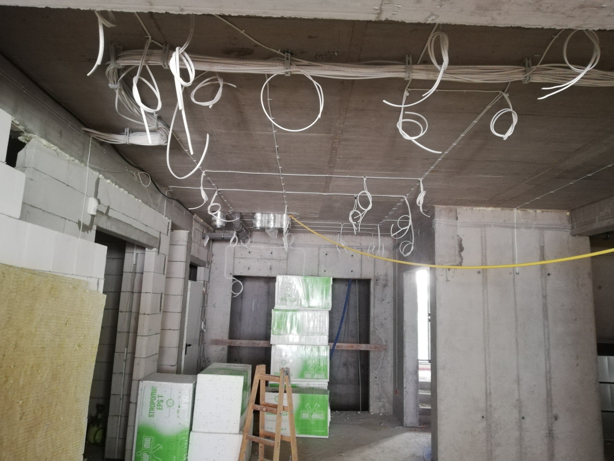 Surowy betonowy strop z widoczną instalacją elektryczną w postaci białych kabli zwisających w pętlach, w tle ściany z bloków i ocieplenia, oraz drabina oparta o ścianę z izolacją.