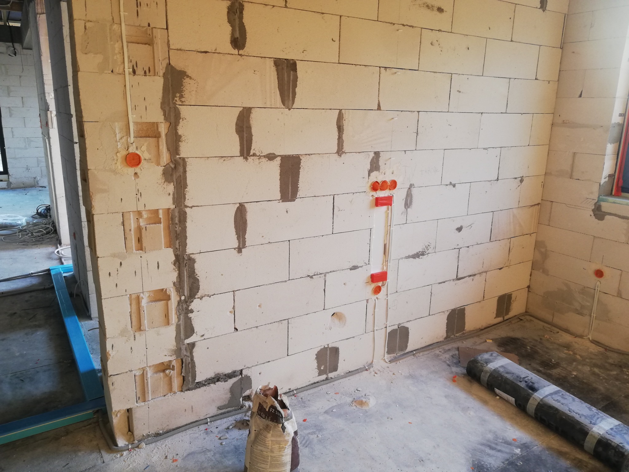Wnętrze w stanie surowym: bloczki z betonu komórkowego z wyżłobieniami pod instalacje elektryczne, widoczne puszki instalacyjne i przewody w peszlach, na podłodze worek z zaprawą i rolka izolacji.