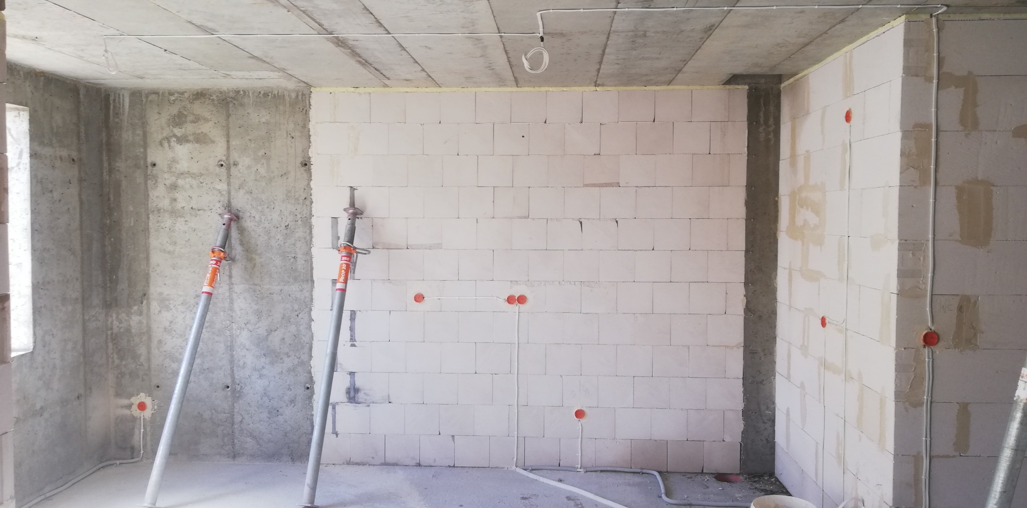 Surowy stan wnętrza z instalacją elektryczną w trakcie montażu, widoczne puszki elektryczne w ścianie z betonu komórkowego i kable prowadzone po powierzchni, podparte stemple budowlane.