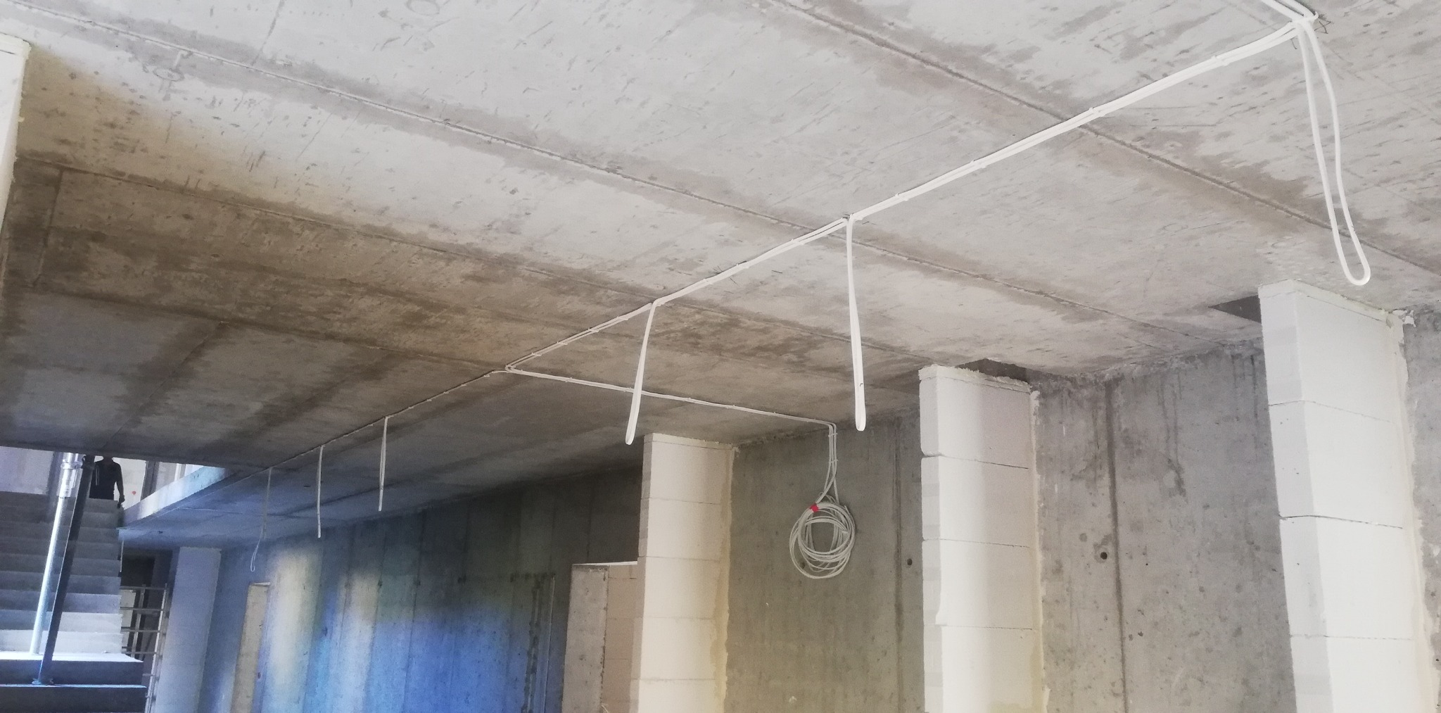Surowy betonowy strop z poprowadzonymi białymi przewodami elektrycznymi, zwisający kabel w pętli, ściany z betonu komórkowego w stanie surowym.