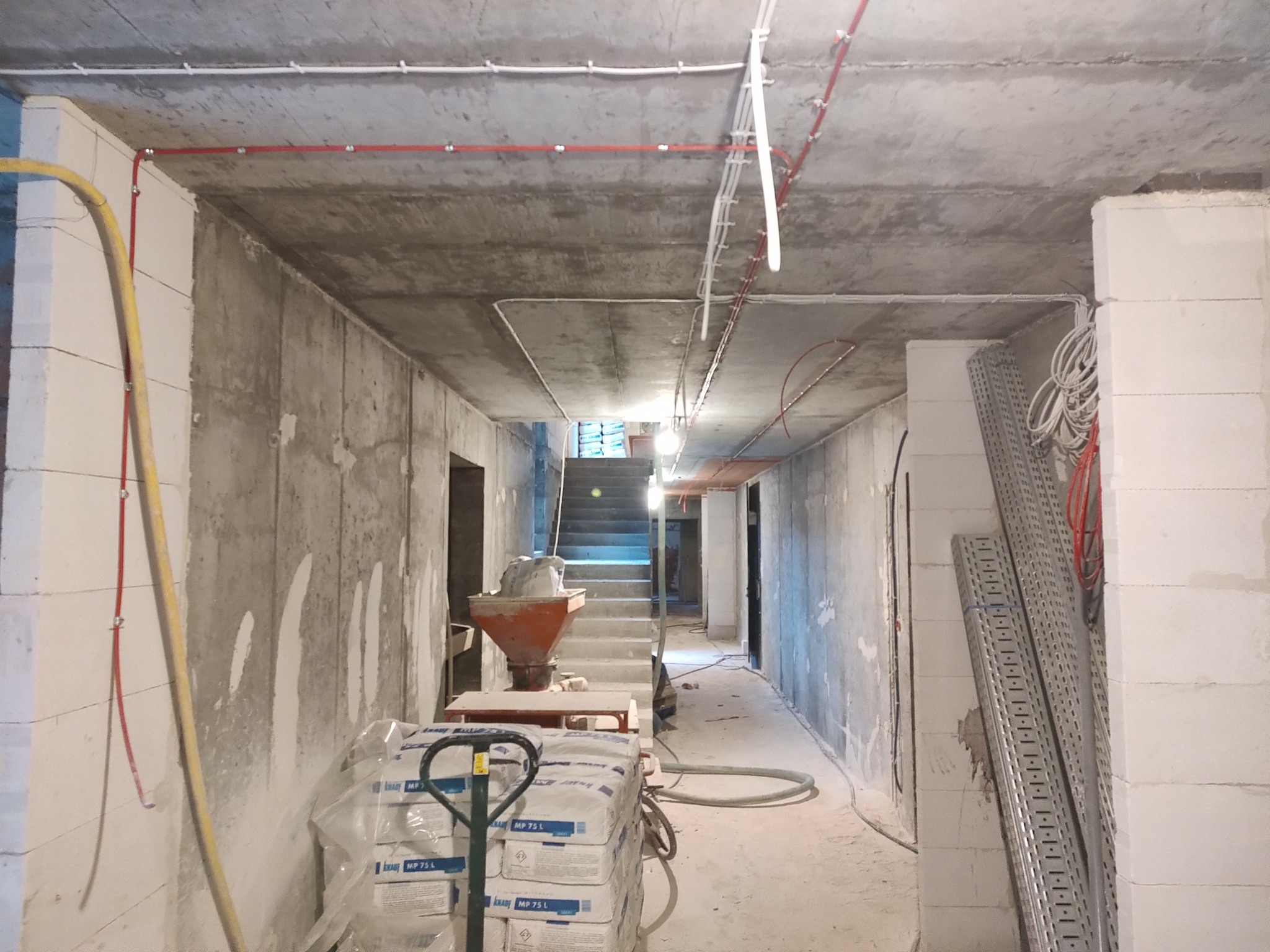 Wnętrze w stanie surowym z widocznymi przewodami elektrycznymi pod sufitem, workami z zaprawą i drabinkami kablowymi, ukazujące etap prac instalacyjnych.