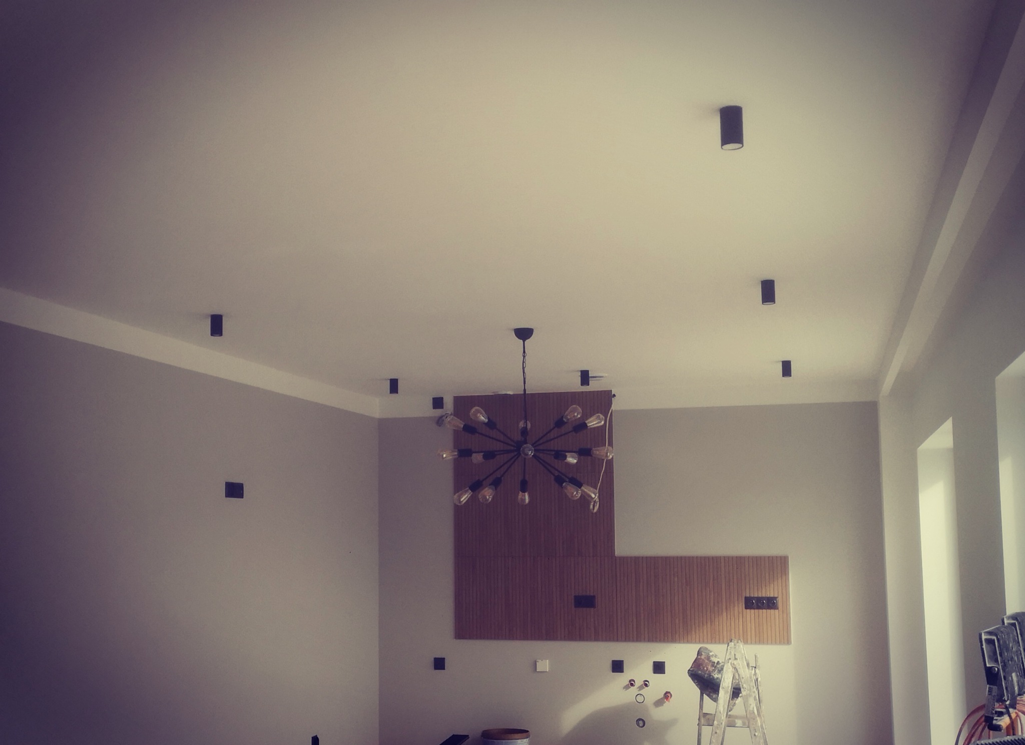 Wnętrze z zamontowanymi punktowymi lampami sufitowymi i nowoczesnym żyrandolem, widoczne przygotowania do dalszych prac wykończeniowych.