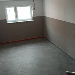 Pusty pokój po remoncie z szarymi płytkami podłogowymi, ozdobną tapetą do połowy ściany i wystającymi kablami z sufitu.
