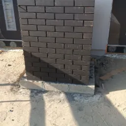 Nowo wymurowany, ciemnobrązowy filar z cegły klinkierowej na betonowej podstawie, przy elewacji budynku w trakcie budowy, z widocznym piaskiem i elementami budowlanymi.