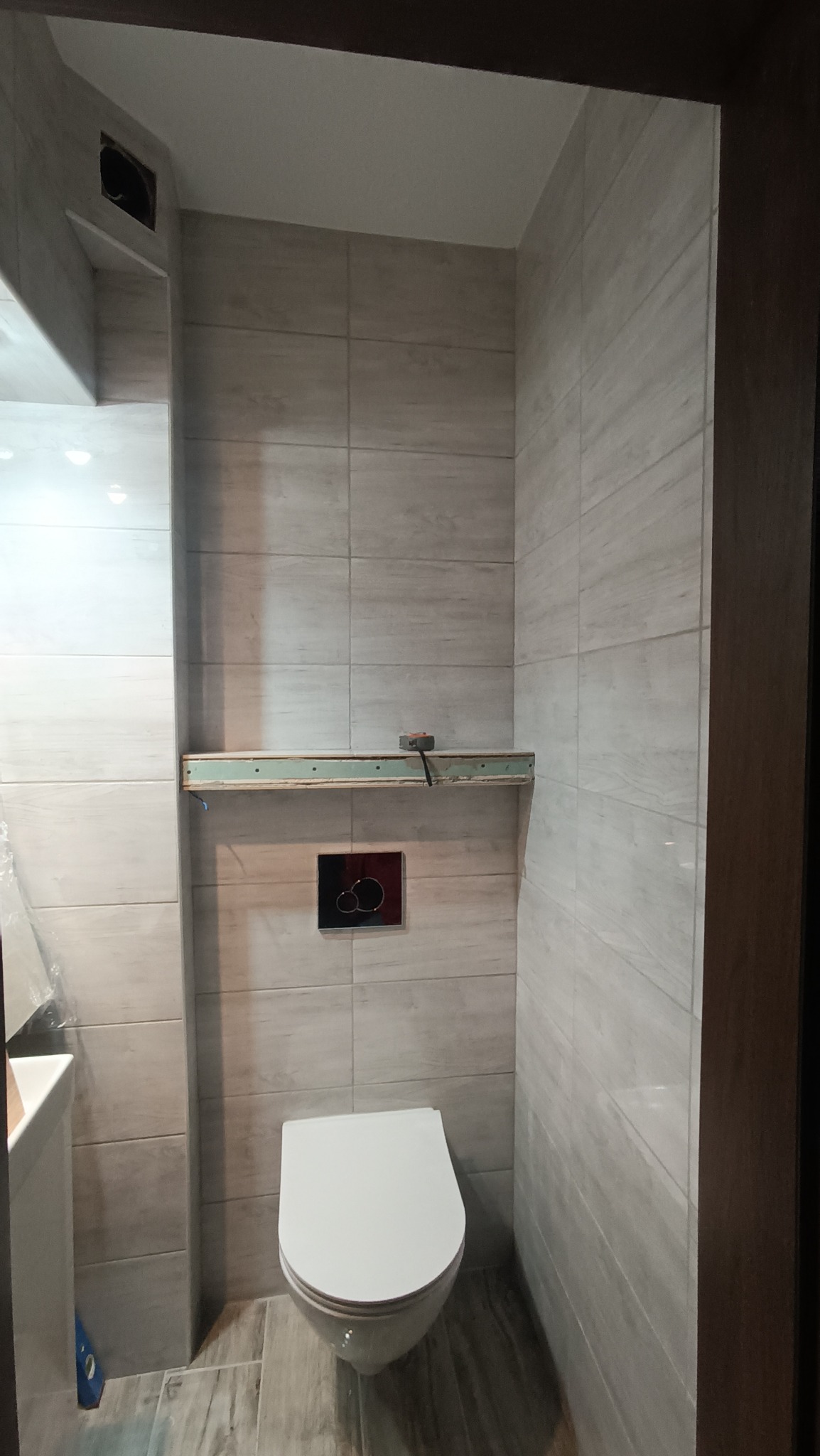 Wnętrze łazienki w trakcie remontu, widoczna podwieszana toaleta, płytki imitujące drewno na ścianach, stelaż pod półkę z widocznymi przewodami i puszką instalacyjną.